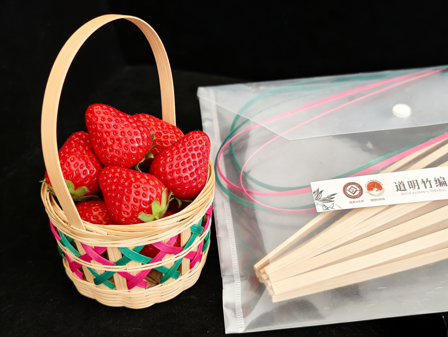 Bamboo lattice basket filled with fresh strawberries next to materials kit with natural bamboo strips on black background