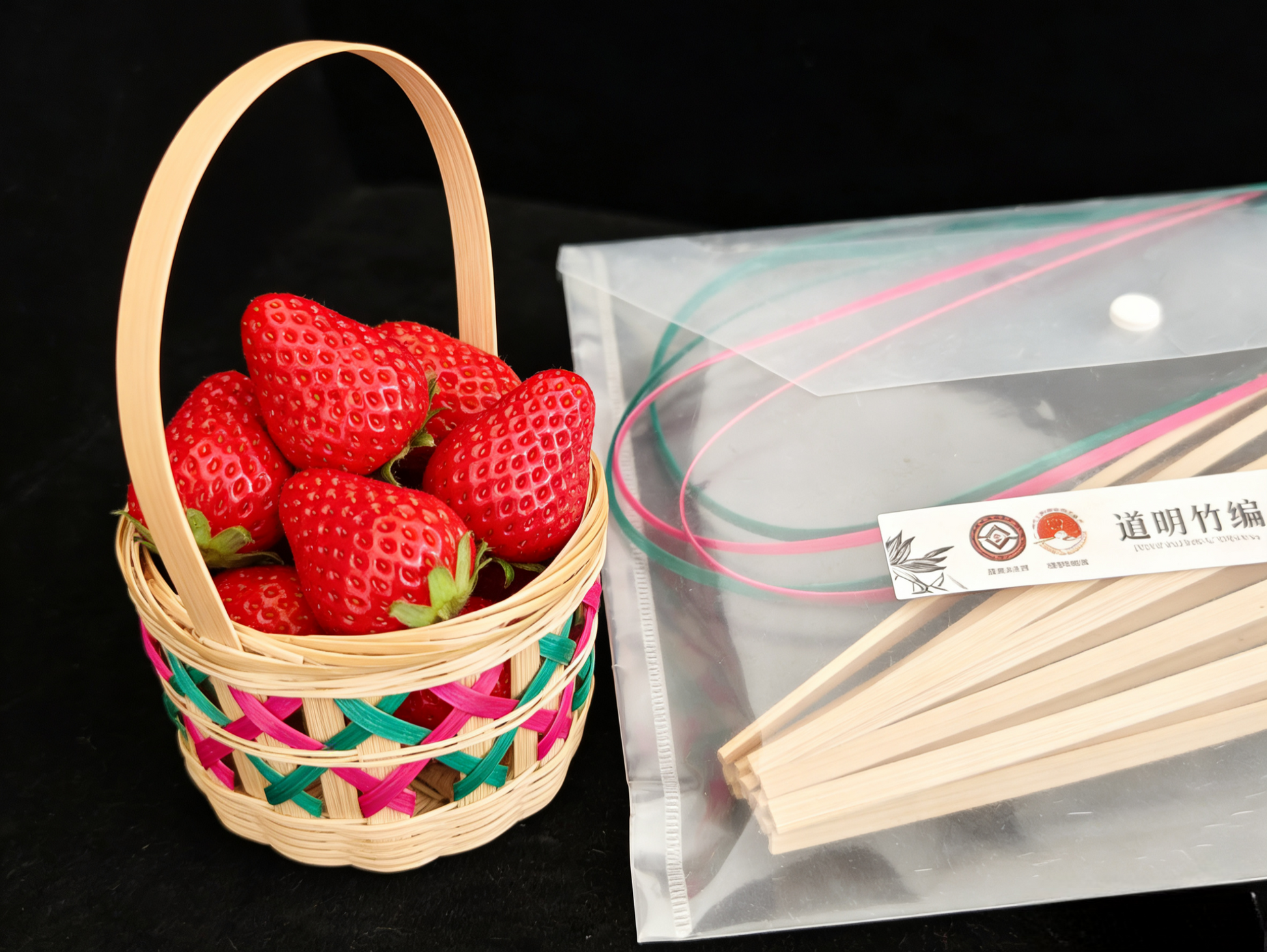 Bamboo lattice basket filled with fresh strawberries next to materials kit with natural bamboo strips on black background