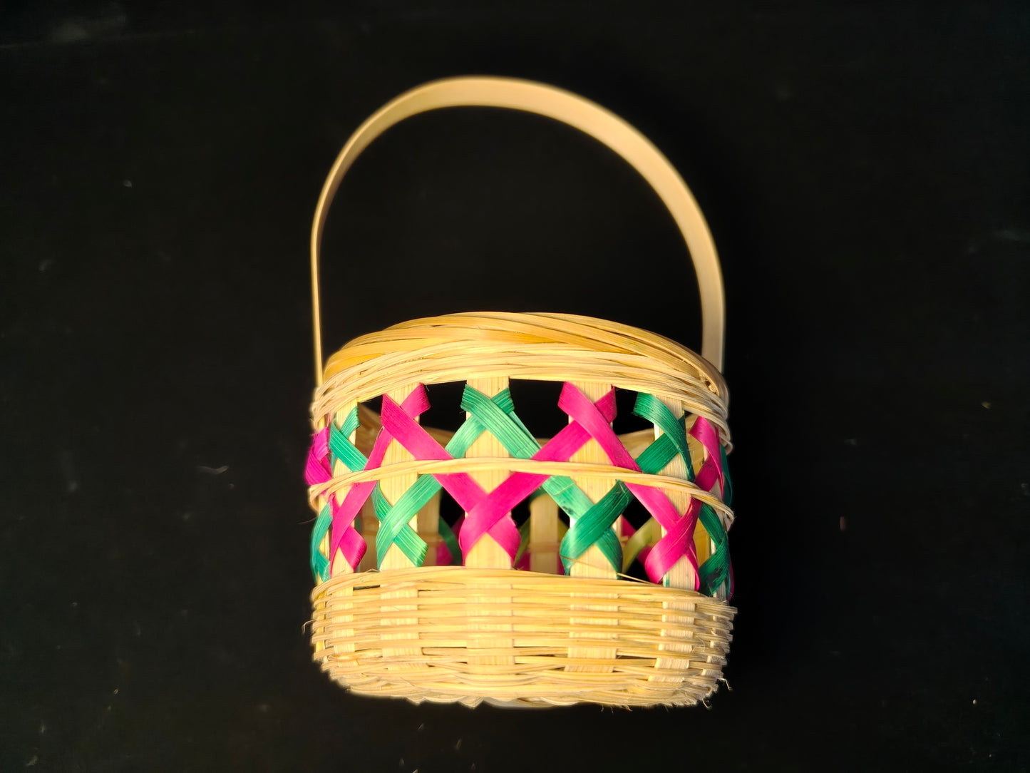Chinese bamboo woven basket with pink and green lattice design and natural handle on black background - side view